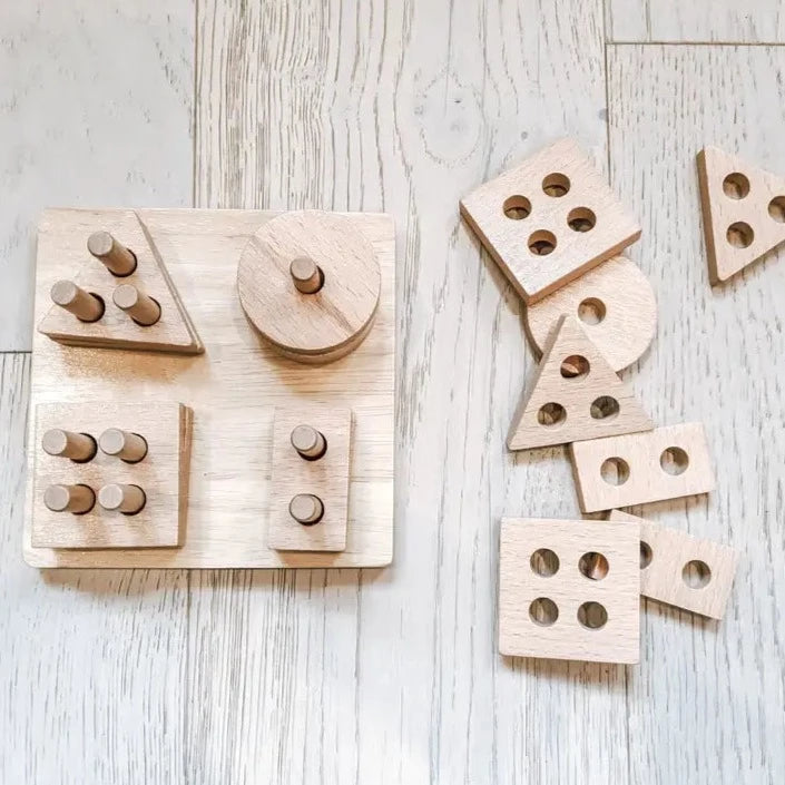 Overhead view of The Shape Sorter by Welly & Bee on a wooden floor. The shape sorter is a square wood base with pegs, andthe shapes are square, triangle, rectangle & circle.