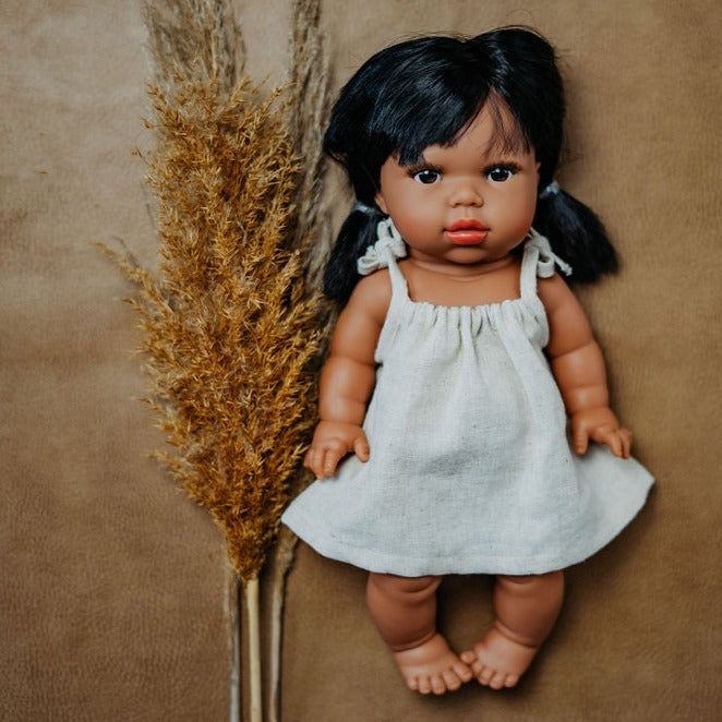 Brown background with pampas grass on the left and the Latika Doll by Minikane, wearing a dress. Latika is an indigenous doll, with dark eyes, and long dark hair.