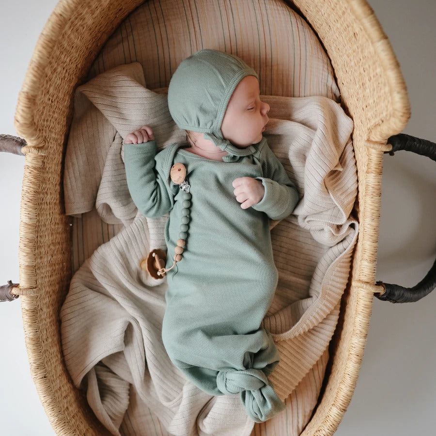 Overhead view of a baby sleeping in a rattan bassinet, wearing a Ribbed Knotted Baby Gown by Mushie.