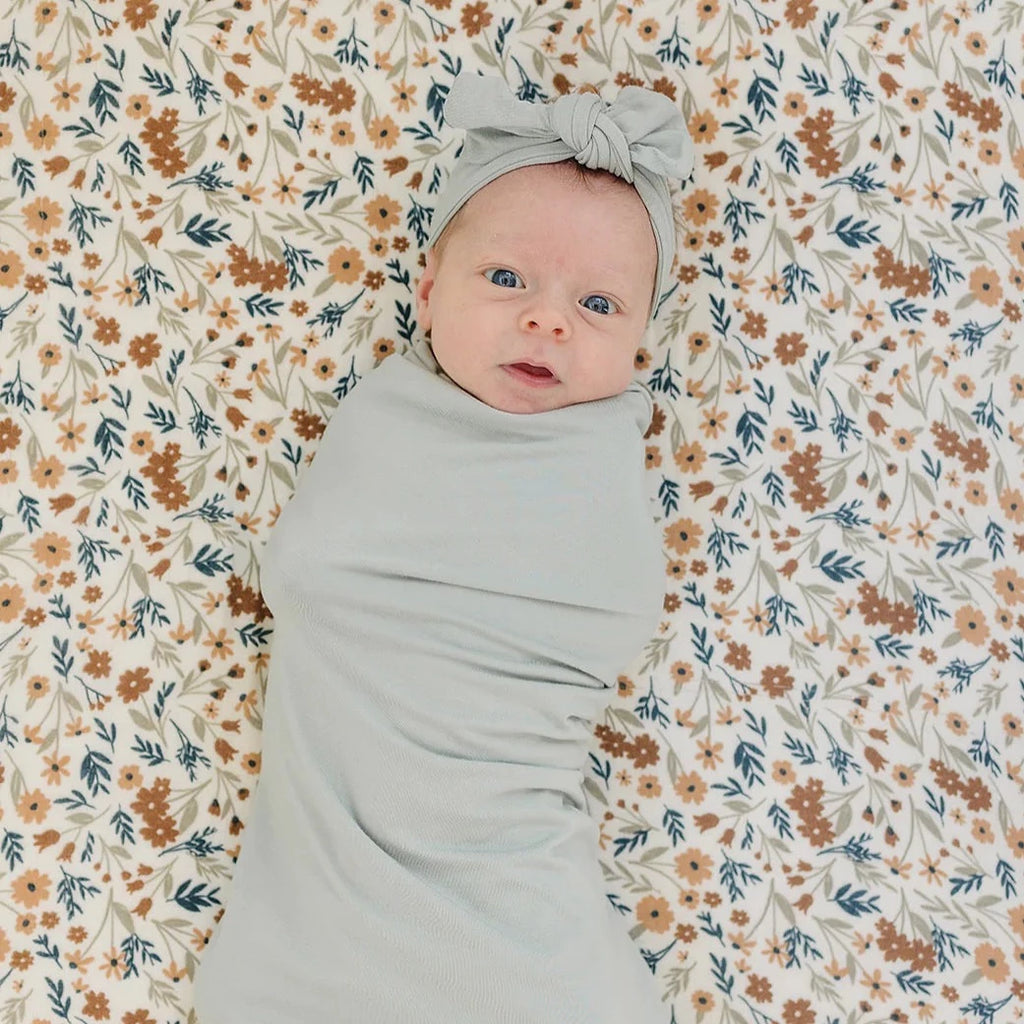 Overhead view of baby laying down wearing a Sage Head Wrap by Mebie Baby. Head wrap is wide, with a knot bow in the front, and is sage.