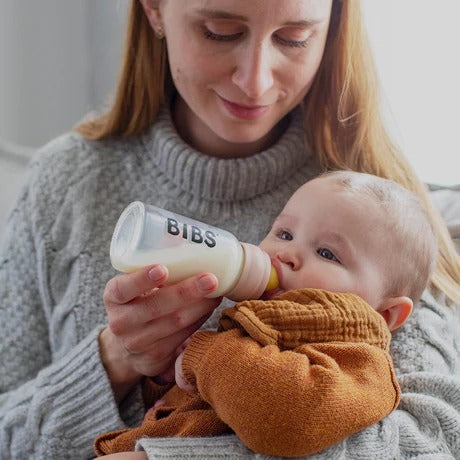 Woman holding baby, feeding them from a glass bottle that says "BIBS" in black with blush colour around bottle rim by Bibs