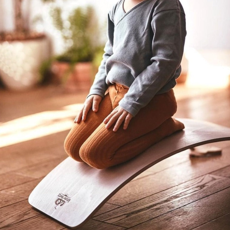 Close up of kid sitting on Lite Natural Kinderboard by Kinderfeets. Kinderboard is a board thats angled in an arch for kids to play with.