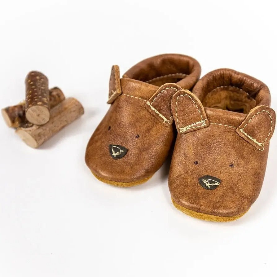 White background with a small stack of logs on the left, and the Russet Bear Leather Moccs by Starry Knight Design. Moccs are a warm brown colour with a bear face, and little bear ears, all on the toe area.