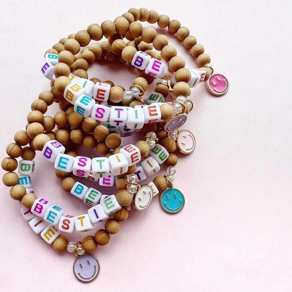 Bestie bracelet by cutieCHEWS. Bracelets have wood beads with bestie on plastic cubed bead with smiley pendant on a cream/pink backdrop.