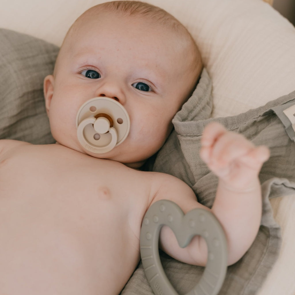 Baby laying down with a sand coloured pacifier in mouth by Bibs