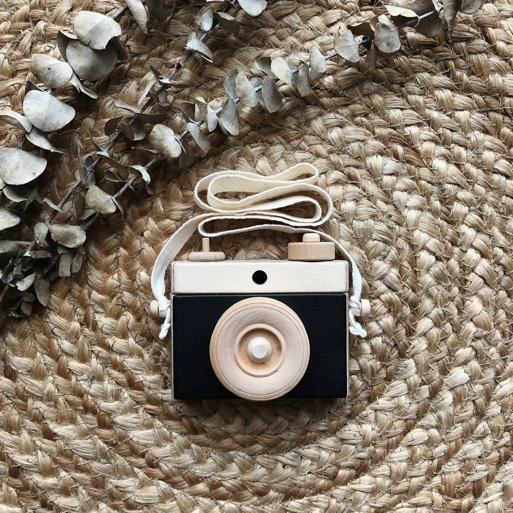 Rattan background with a Wooden Camera in Black by Little Rose & Co in the centre. Camera is wooden with black accents, and a canvas strap.