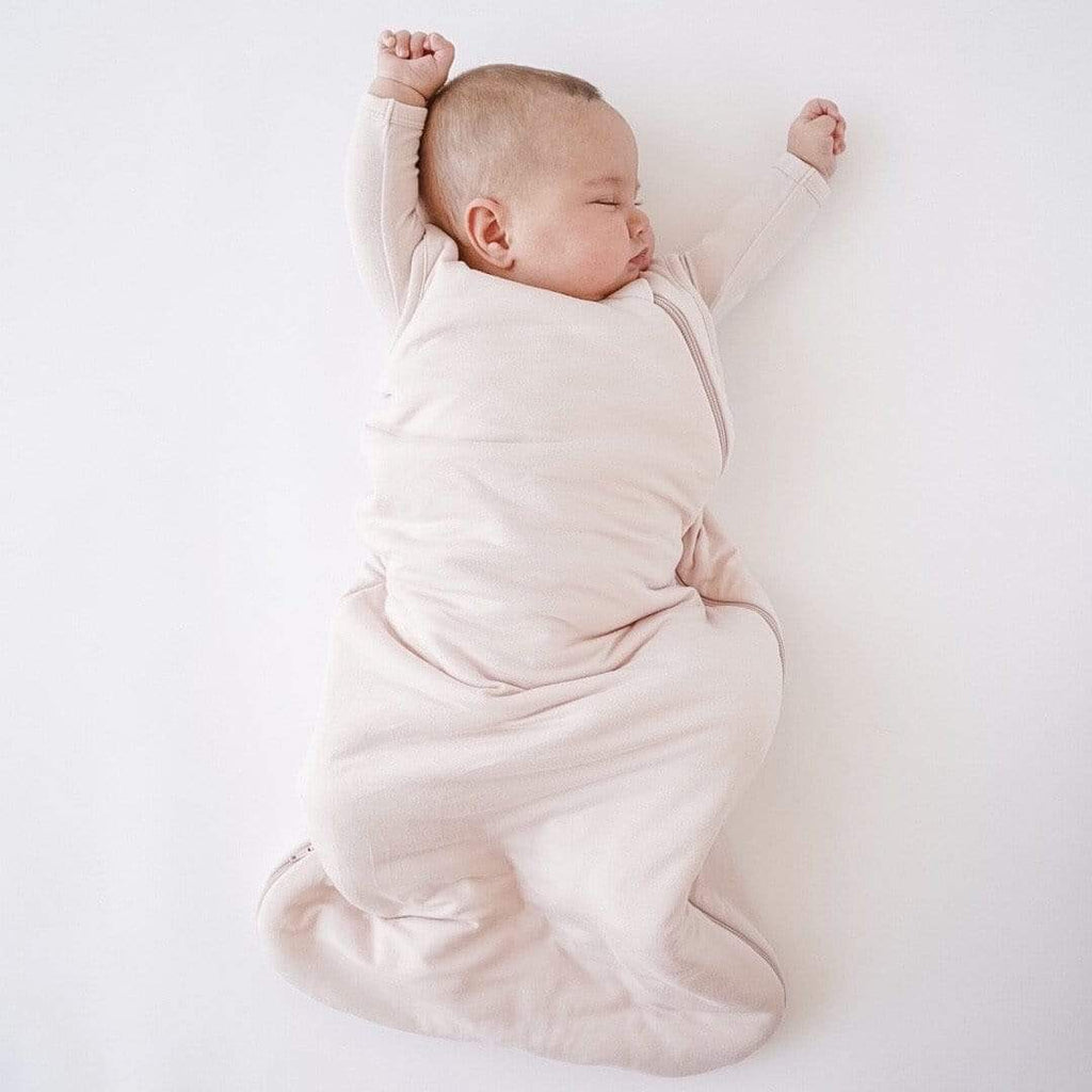 White background with baby laying down, sleeping, wearing the Sleep Bag 1.0 Tog in Blush by Kyte Baby. Sleep bag is a light pink colour with a zipper that does from the upper left shoulder all the way down the side, and to the bottom.