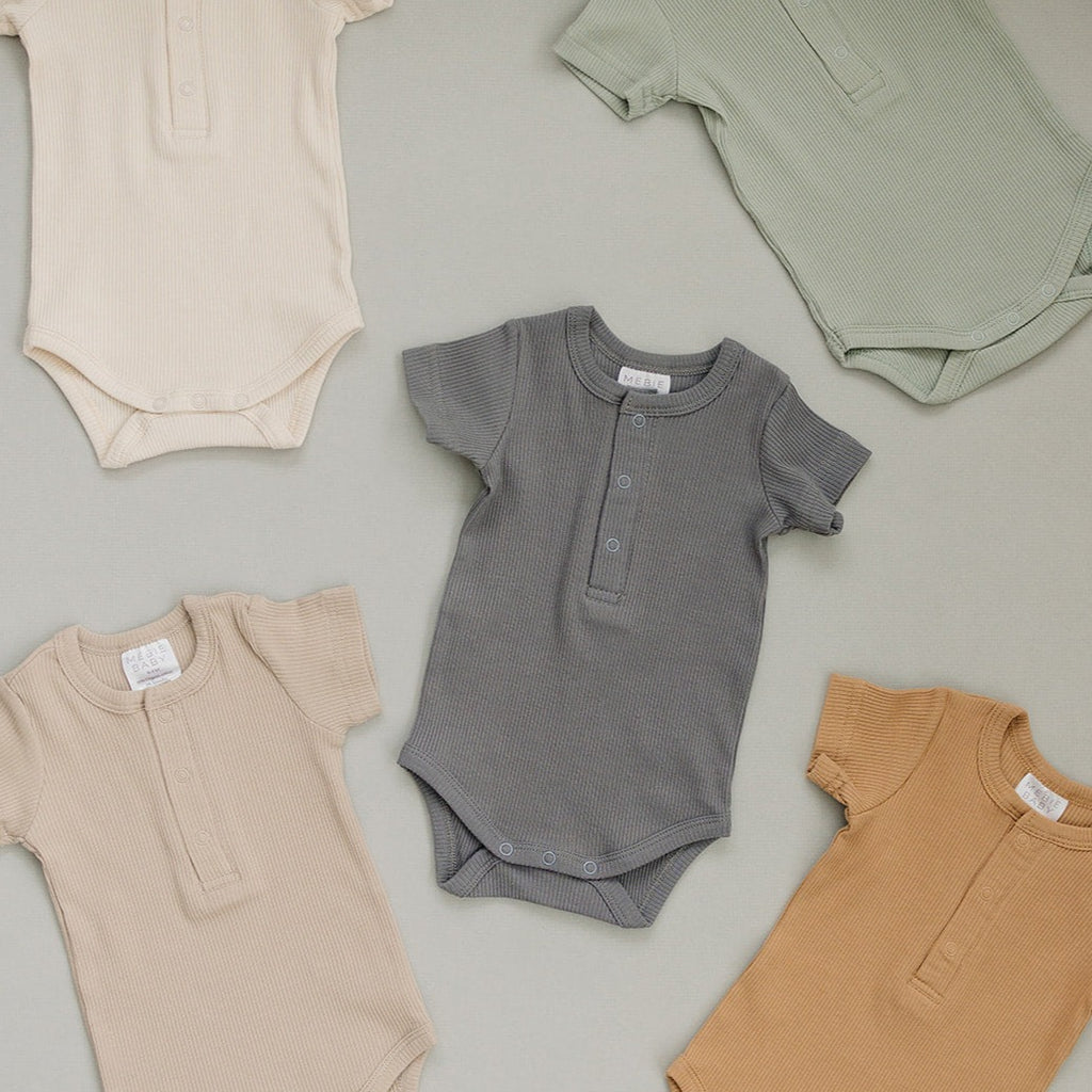 Mebie Baby Organic Bodysuits in multiple colours, laid on a greige surface. 