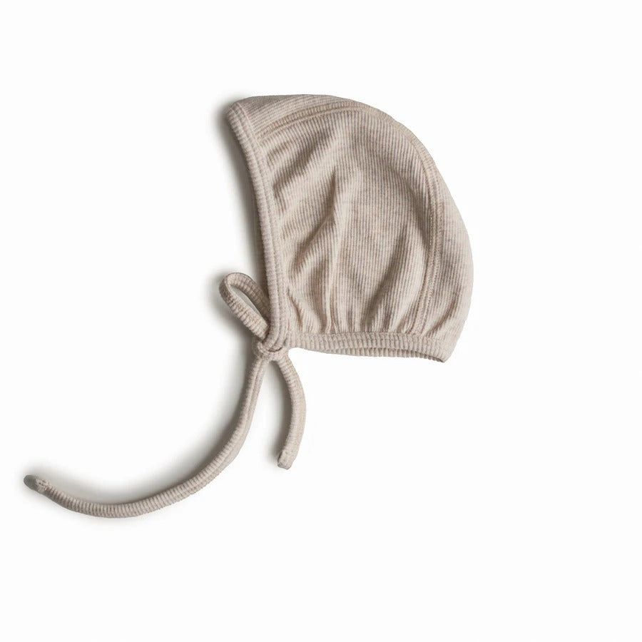 White background with a side view of the Ribbed Baby Bonnet in Beige Melange by Mushie. The bonnet is a ribbed material in an oatmeal colour, that fits snug on the head with a tie for under the chin.
