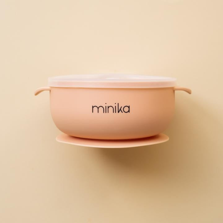 Beige background with a Silicone Bowl with Lid in Blush by Minika. Bowl is blush, has a silicone base to stick to your table, and a clear lid.