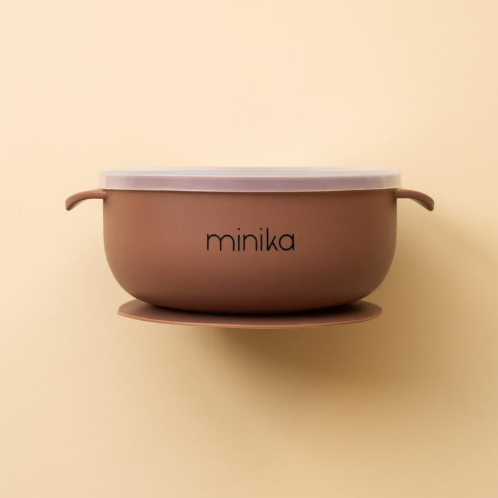 Beige background with a Silicone Bowl with Lid in Cacao by Minika. Bowl is dark brown, has a silicone base to stick to your table, and a clear lid.
