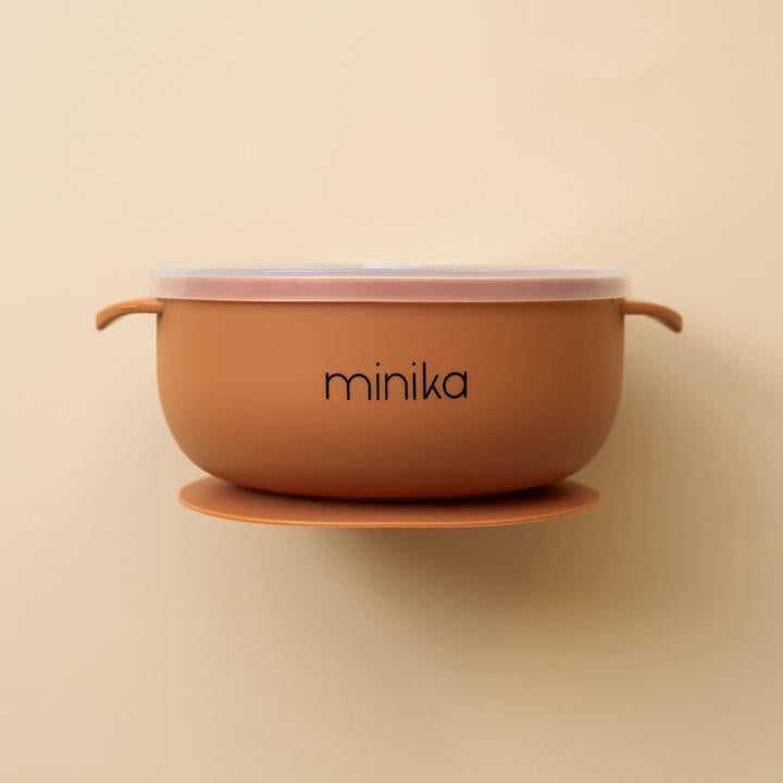 Beige background with a Silicone Bowl with Lid in Ginger by Minika. Bowl is orange/rust, has a silicone base to stick to your table, and a clear lid.