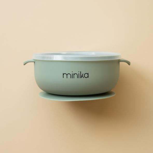 Beige background with a Silicone Bowl with Lid in Sage by Minika. Bowl is sage, has a silicone base to stick to your table, and a clear lid.