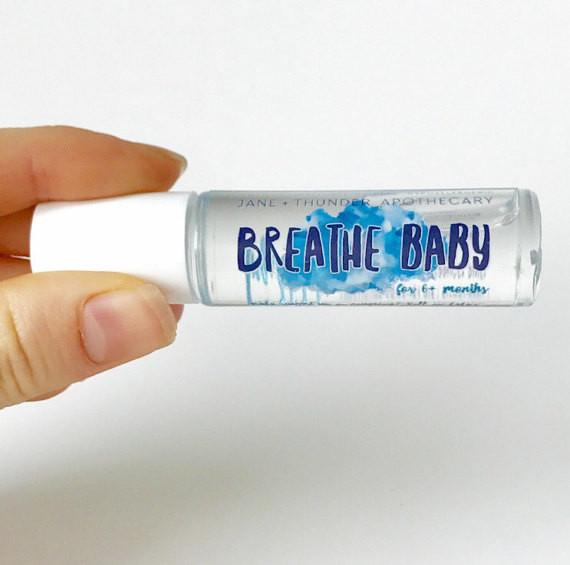 White background with a hand holding the Breathe Baby - Baby Safe Essential Oil by Jane + Thunder. Bottle says "Breathe Baby" in blue font.