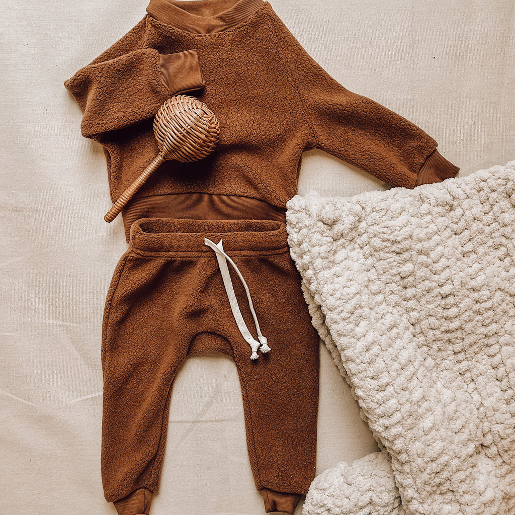 Downtown Brown crew and pants by Petit Nordique, with a rattan baby rattle and a crocheted beige blanket by Petit Nordique, laid on a flat beige surface. 