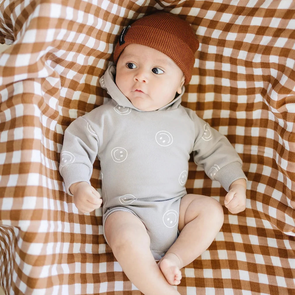Overhead view of a baby boy laying in a bassinet, with a gingham quilt underneath him, wearing the Brown Beanie by Mebie Baby.