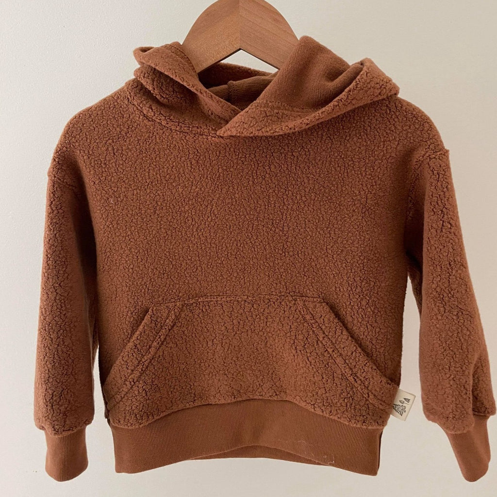 Downtown Brown Sherpa Hoodie by Petit Nordique, hanging on a wooden hanger with a white back drop. 