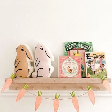 Floating shelf with a carrot banner, easter books and 2 Bunny Pillows by Imani Collective. The first bunny is tan, and the second is lavender.