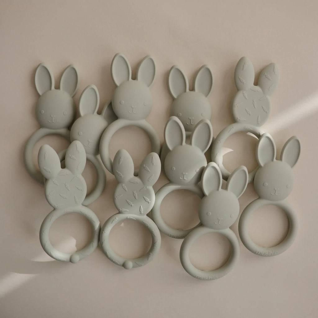 Grey/Beige background with a pile of Bunny Teethers by Mushie. Teethers are silicone sage green, with a round handle, and a bunny head with ears on the top. The backside has little engraved carrots, for texture while teething.