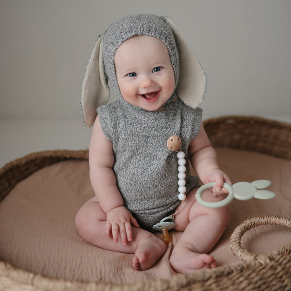 Grey background with a rattan bassinet, and a baby dressed as a bunny holding the Bunny Teether by Mushie.