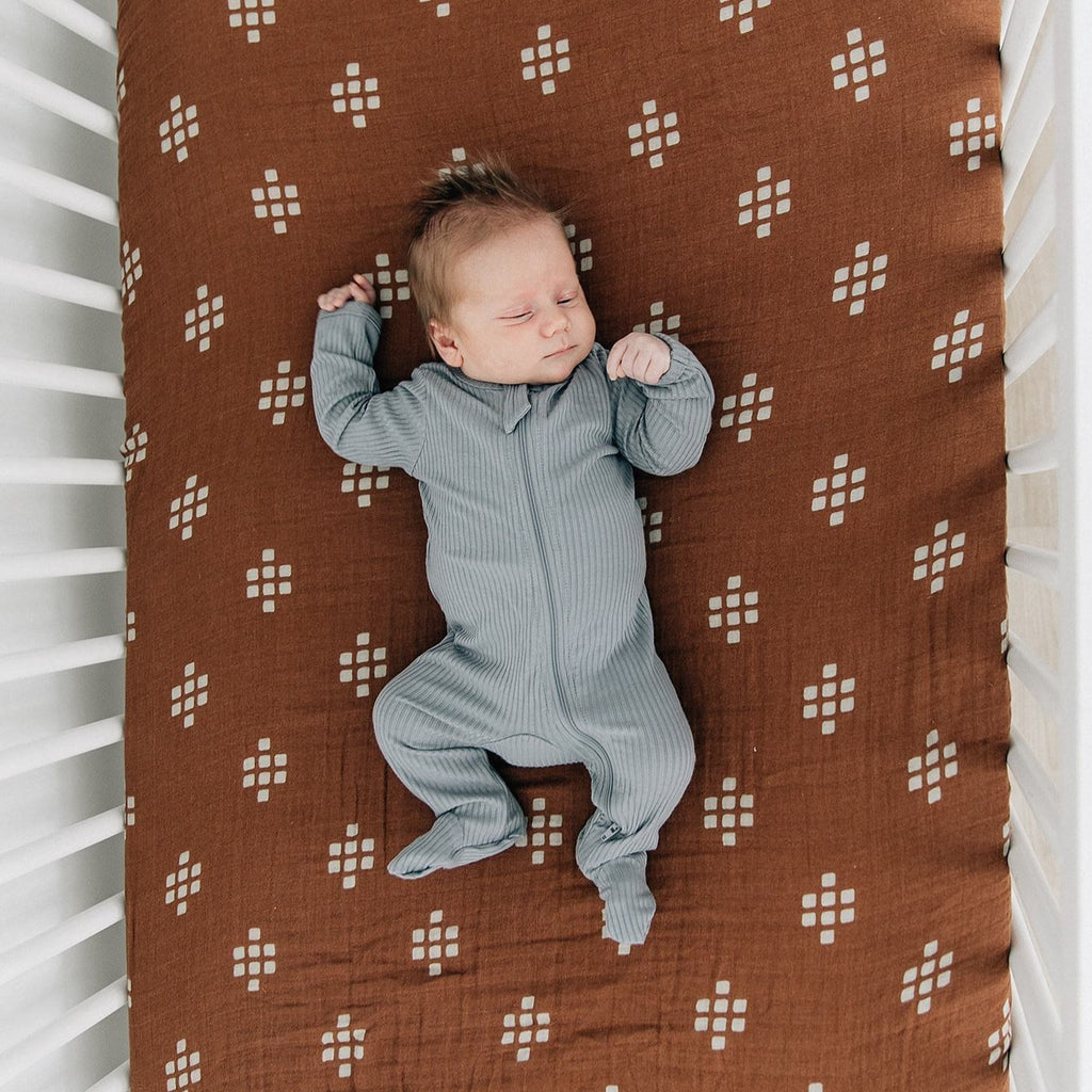 Overhead view of a baby sleeping on a bed with a Chestnut Textiles Crib sheet  by Mebie Baby on the mattress. Crib sheet is dark brown with cream square pattern.
