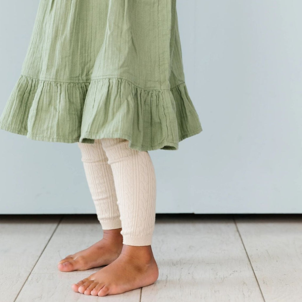 Close up of little girl standing in a dress, wearing Cable Knit Footless Tights in Vanilla by Little Stocking Co. 