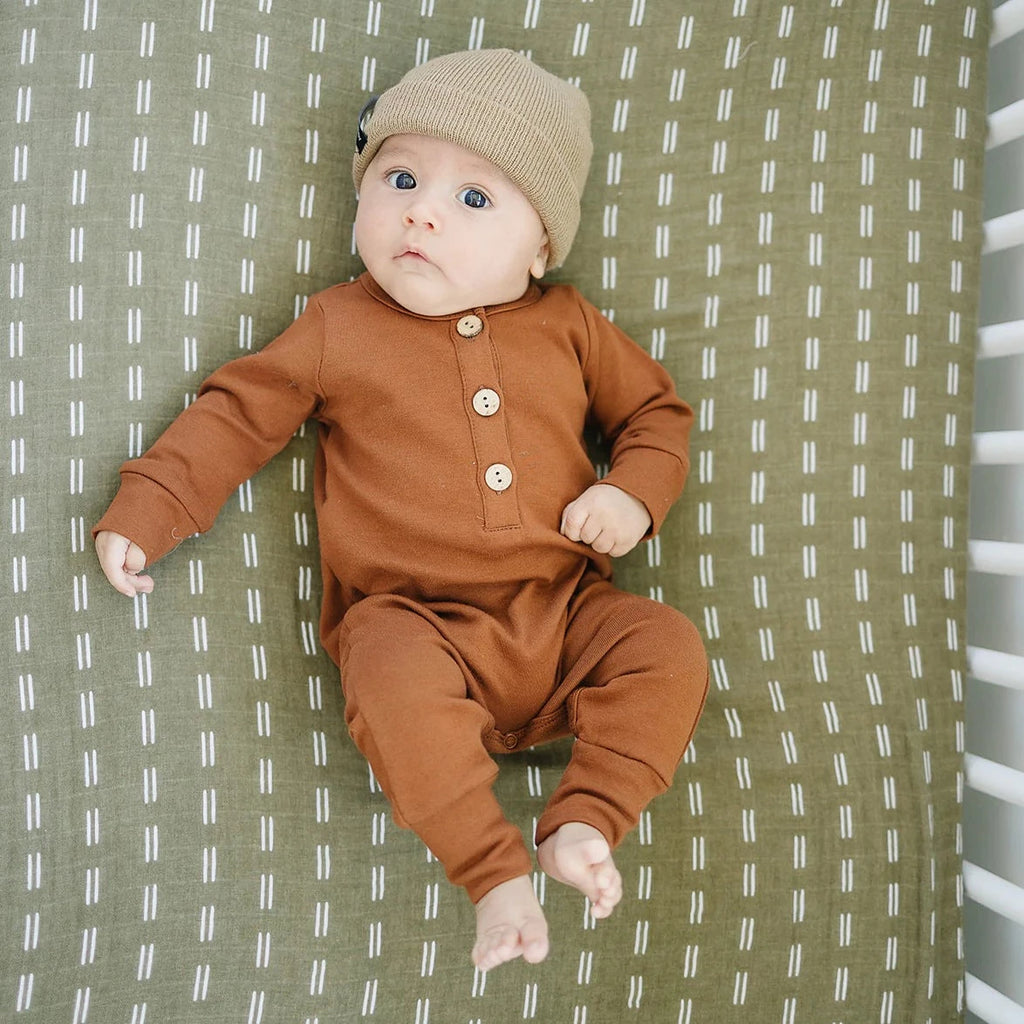 Overhead view of a baby boy laying on the Olive Strokes Crib Sheet, wearing a cafe Au Lait Beanie by Mebie Baby. 