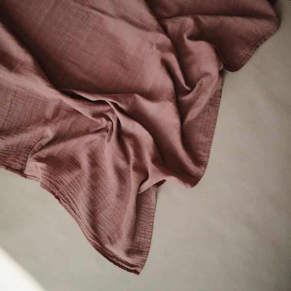 Cream background with the Muslin Swaddle Blanket Organic Cotton in Cedarby Mushie. This swaddle is a burgundy/brown colour.