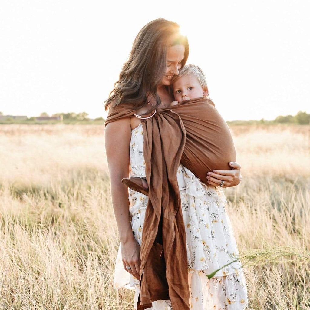 Woman standing outside in a field, the sun behind her, she's looking down at baby in Ring Sling in Cedar by Kyte Baby. Ring sling is a warm brown colour, with a rose gold ring.