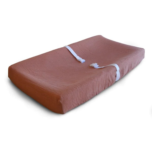 White background with a changing pad, and an Extra Soft Muslin Changing Pad Cover in Cedar by Mushie on it. Cedar is a burgundy colour.