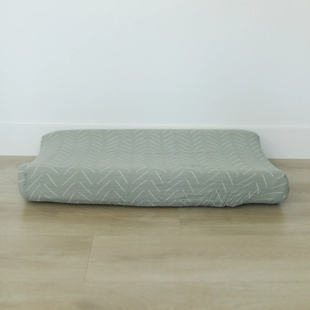 White wall with a light wood floor, and the Desert Sage Changing Pad Cover by Mebie Baby on a change pad. This cover is muslin in a soft sage colour, with white lines.