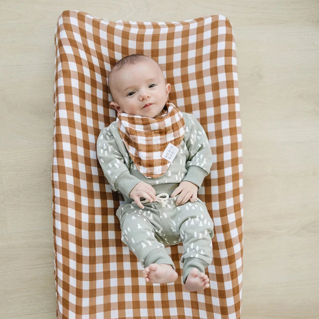Wood background with an overhead view of a baby laying on a change pad with the Gingham Changing Pad Cover by Mebie Baby on it.
