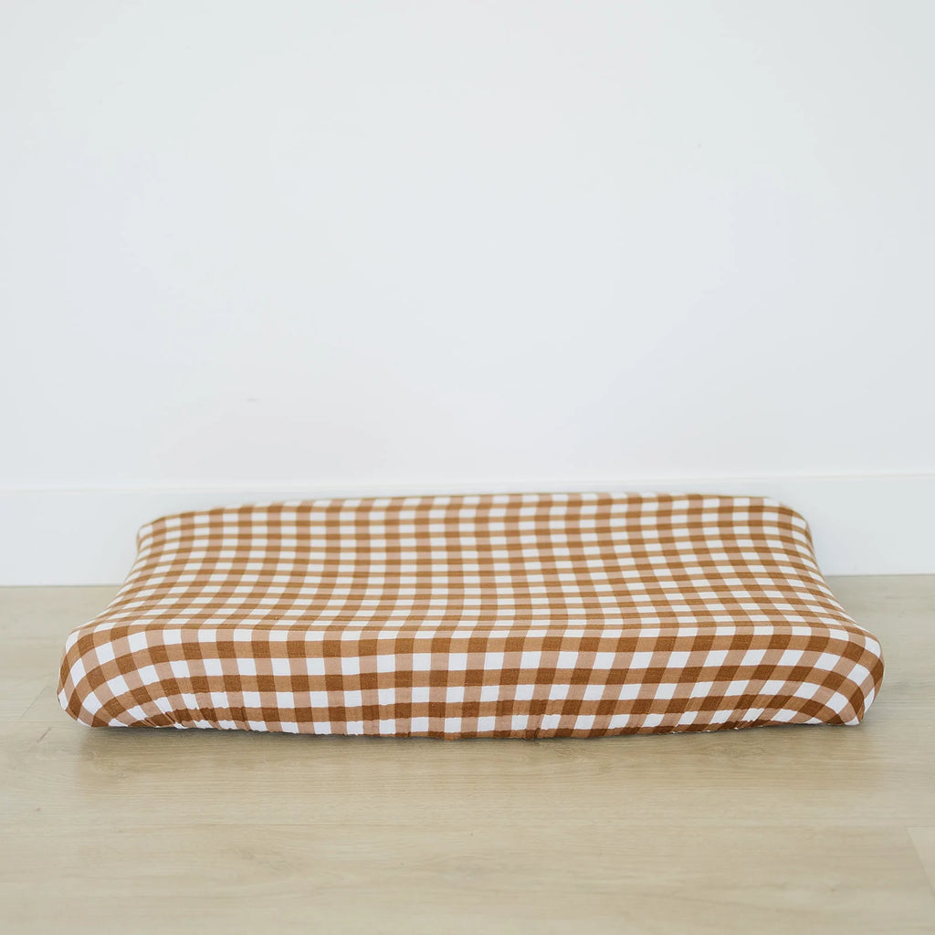 White background with a light wood floor, and a changing pad with the Gingham Changing Pad Cover by Mebie Baby on it. This cover is a white and dark brown gingham pattern.
