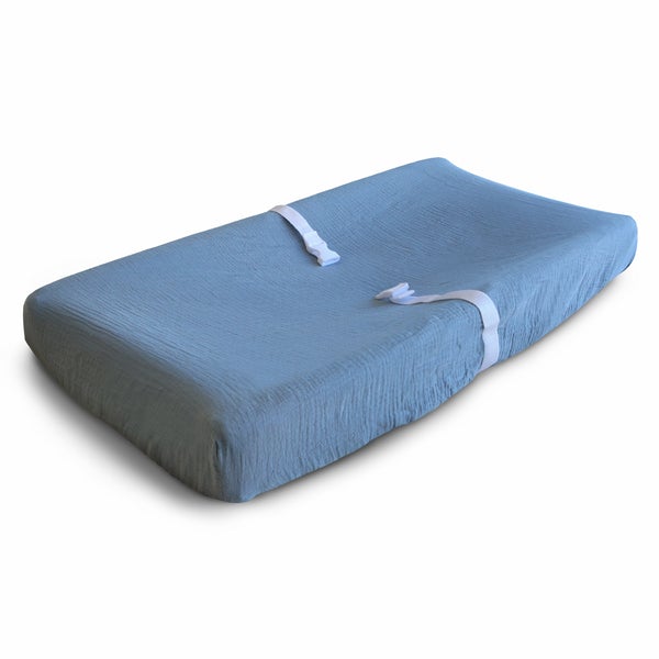White background with a changing pad, and an Extra Soft Muslin Changing Pad Cover in Trade Winds by Mushie on it. Trade Winds is a medium cool blue.