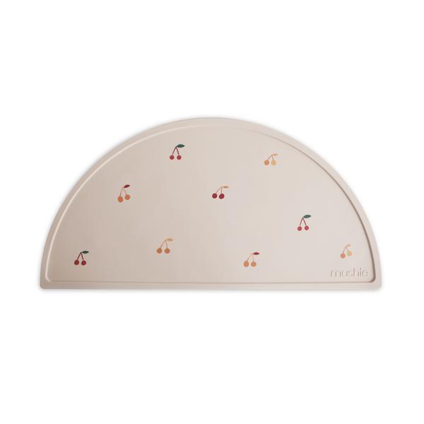 White background with Silicone Place Mat in Cherries by Mushie. Placemat is beige with red and orange cherries, made of silicone, and in a semicircle.