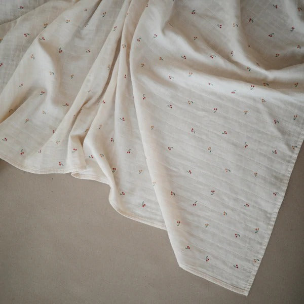 Cream background with the Muslin Swaddle Blanket Organic Cotton in Cherries by Mushie. This swaddle is white with little red and orange cherries all over.