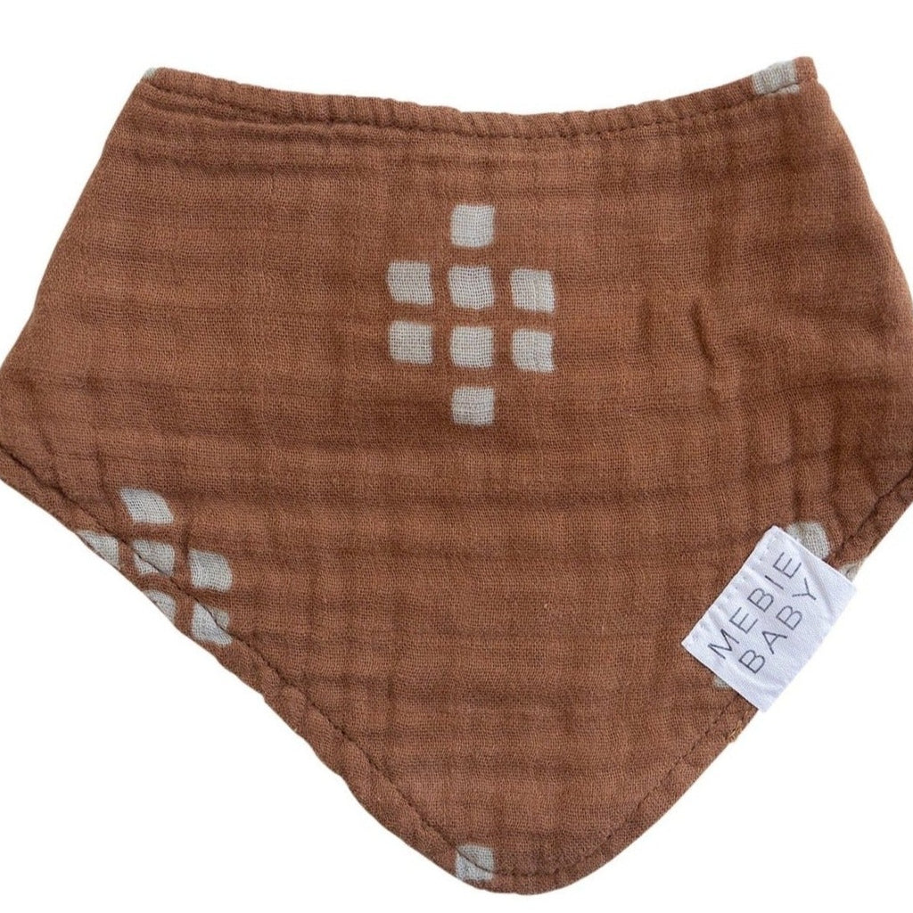White background with Chestnut Textiles Bib by Mebie Baby. Triangle bib is dark brown with cream square pattern all over, and a small white tag that says "MEBIE BABY".