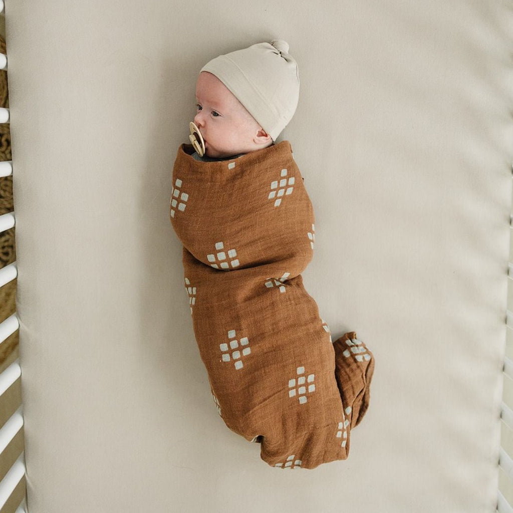 Overhead view of baby laying in crib, wrapped in a Chestnut Textiles Muslin Swaddle by Mebie Baby, Swaddle is dark brown with cream square print.