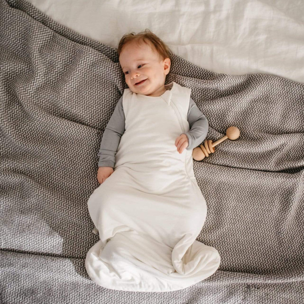 Overhead view of baby laying down, smiling, wearing a Sleep bag 1.0 Tog in Cloud by Kyte Baby. Sleep Bag is white with a side zipper going all the way down.
