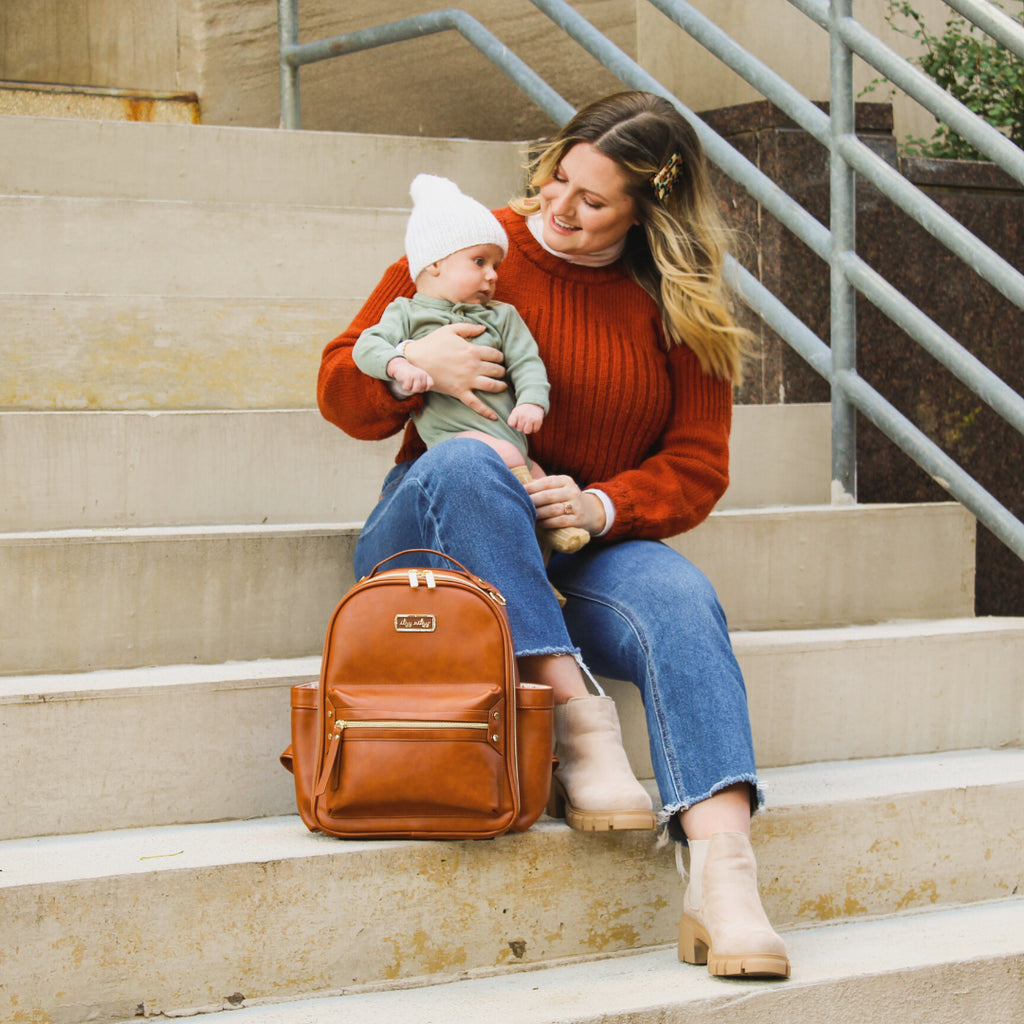 Woman sitting on concrete steps, baby on her lap with the Itzy Mini™ Diaper Bag Backpack in Cognac by Itzy Ritzy beside her. Bag is cognac colour, with gold zippers and a gold tag.