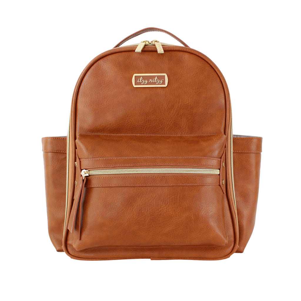 White background with Itzy Mini™ Diaper Bag Backpack in Cognac by Itzy Ritzy. Bag is cognac/rust colour, with gold zippers, and a gold tag that says "itzy ritzy".