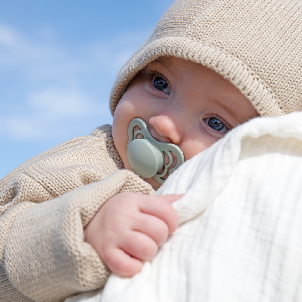 Close up of a baby being held outside, with a bonnet on, and a bright blue sky behind, with the Couture Size 1 Pacifier in Sage by Bibs in their mouth.