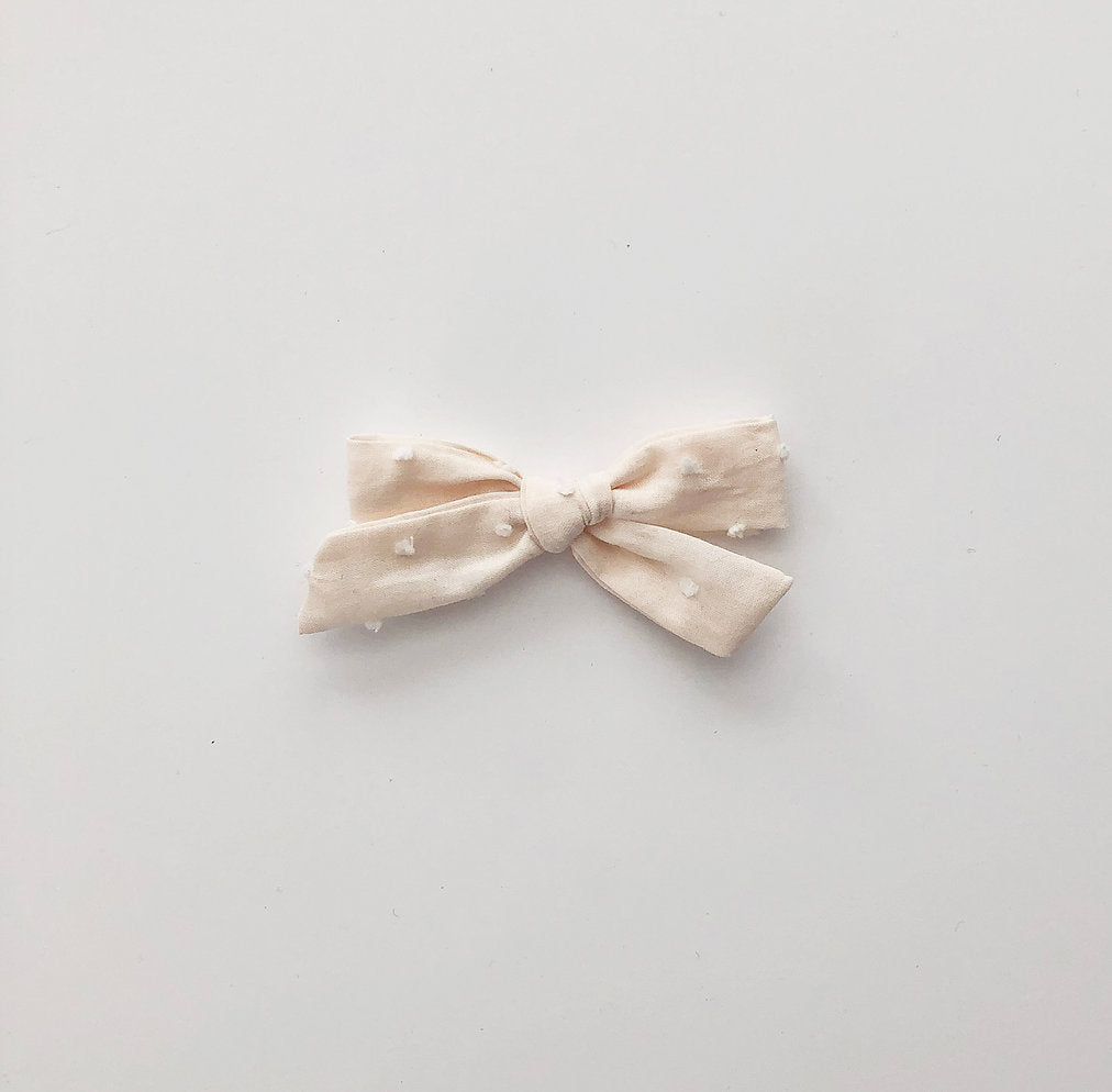 White background with the Cream Swiss Dot School Girl Bow by Wild and Free Design Co. Bow is a pale cream colour, it's thin, and has white swiss dots all over.