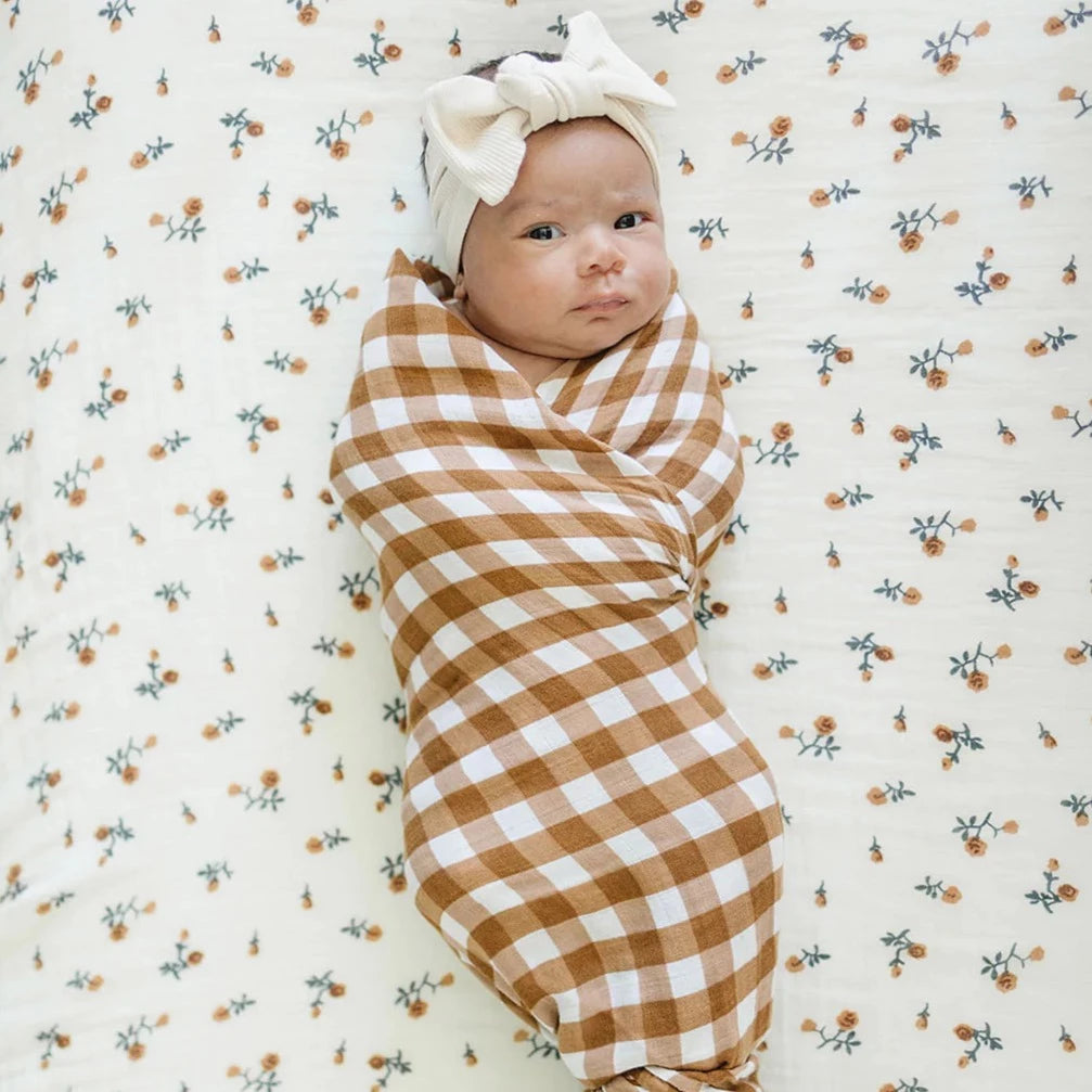 Overhead view of a baby laying on a mattress with the Cream Floral Crib Sheet by Mebie Baby on it, swaddled in a Gingham print, and a Headwrap on. Crib sheet is a light cream colour with rust flowers with dark leaves.