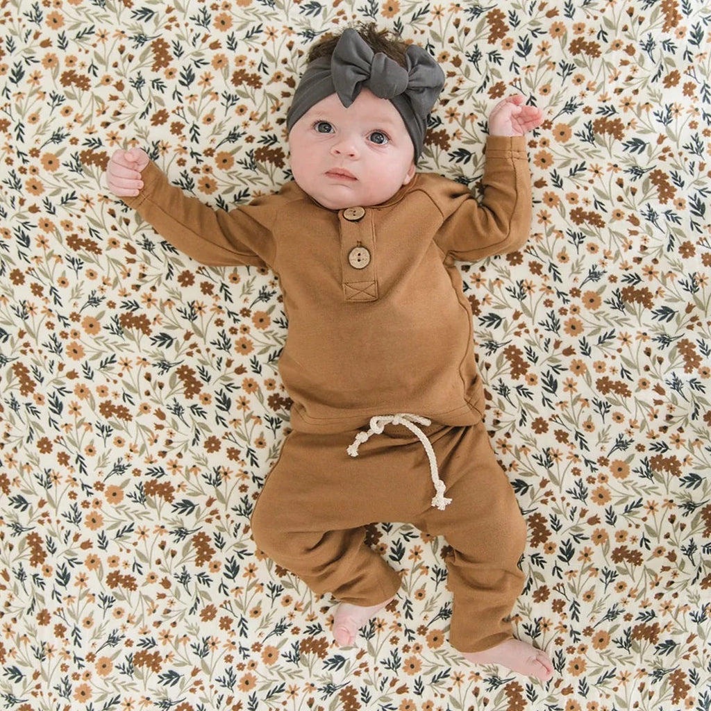 Overhead view of a baby laying down on the Harvest Floral Crib Sheet by Mebie Baby. Crib sheet is a cream/white with a pattern of flowers all over, in rust, blue, and beige.