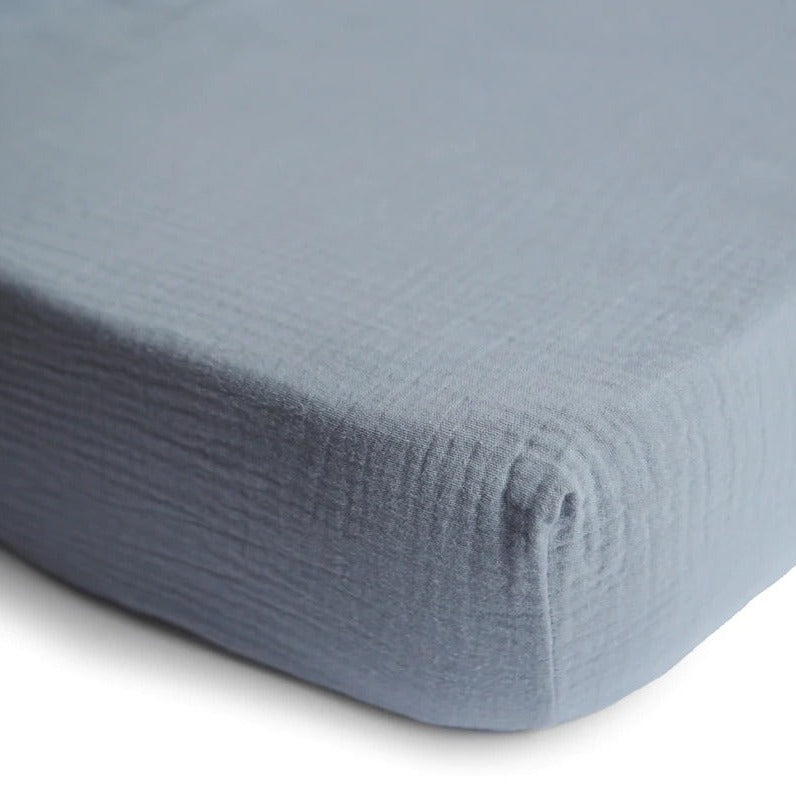 White background with a close up of the corner of a mattress with the Extra Soft Muslin Crib Sheet in Trade Winds by Mushie on it. Trade Winds is a cool grey blue.