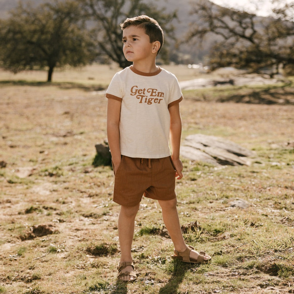 Model wearing Bermuda Short in Camel by Rylee and Cru, while standing in a grass field with trees in the background. 