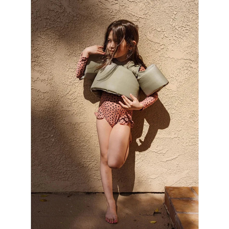 Model wearing an orange and black bathing suit and sage green floaties by Current Tyed. Model is leaning against a stucco beige wall, with a step to the right of her. 