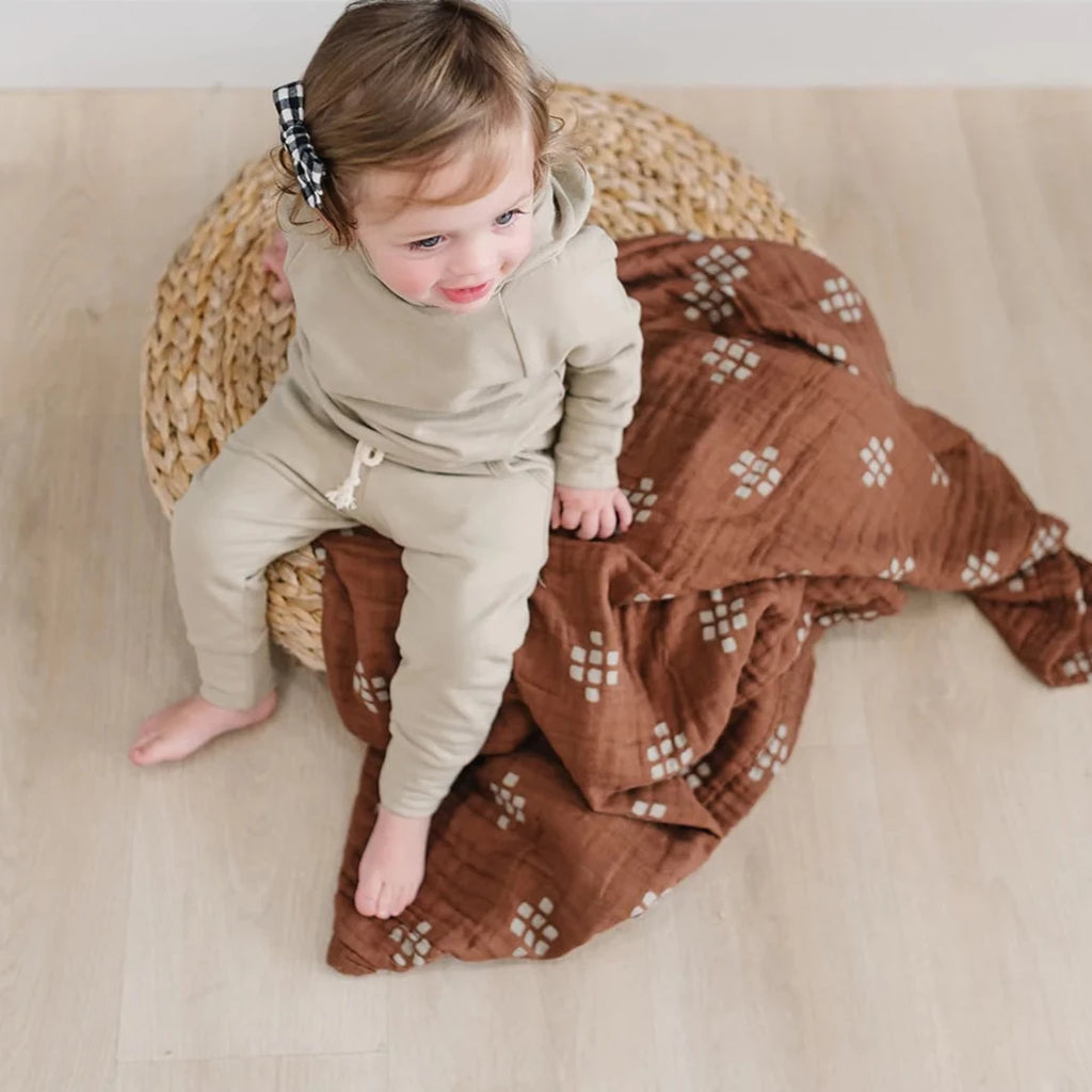 Overhead view of little girl sitting on a rattan ottoman, with a Chestnut Textile Muslin Quilt by Mebie Baby. Quilt is dark brown with cream/white square pattern.