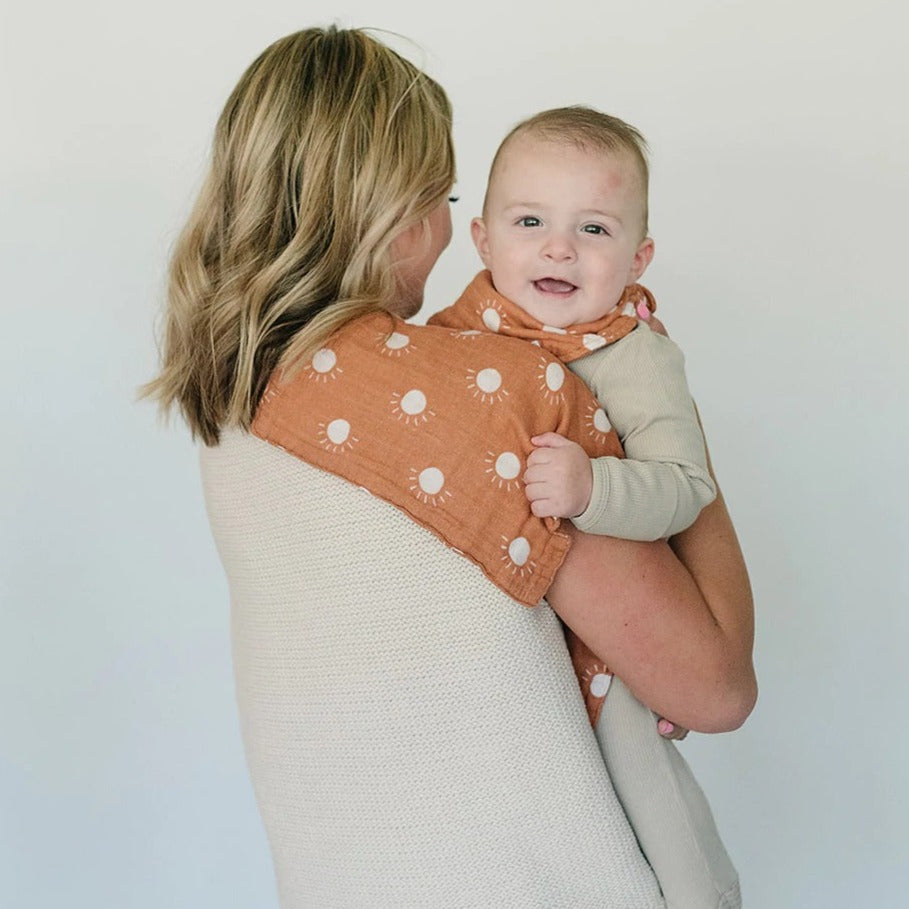 White background with a mama holding her baby, back to the camera, and a Sunshine Burp Cloth by Mebie Baby draped over her shoulder. Burp cloth is orange with white suns all over.